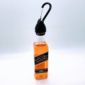 May include: A miniature bottle of Johnnie Walker Black Label Blended Scotch Whisky with a black carabiner attached to the top. The bottle is clear and filled with amber liquid. The label is black with gold lettering and the words "Johnnie Walker Black Label Blended Scotch Whisky" are printed on the label.