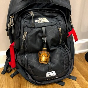 May include: A black The North Face Surge II backpack with red straps and a small bottle of Crown Royal whiskey attached to the front zipper.
