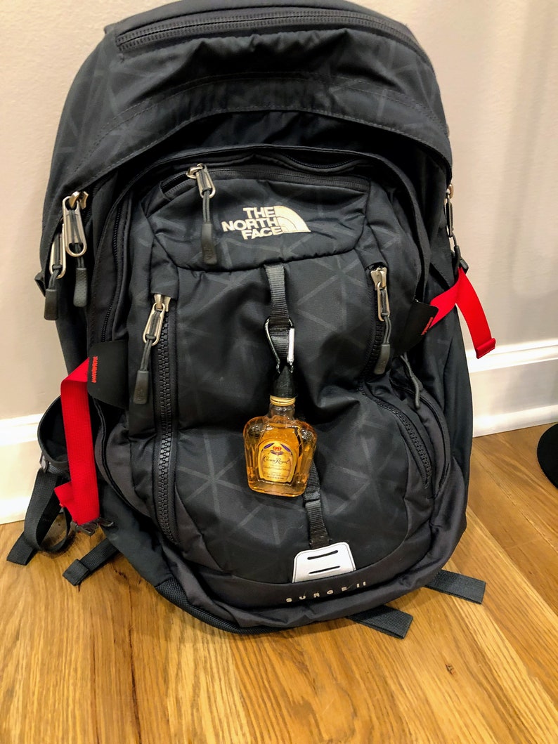 May include: A black The North Face Surge II backpack with red straps and a small bottle of Crown Royal whiskey attached to the front zipper.
