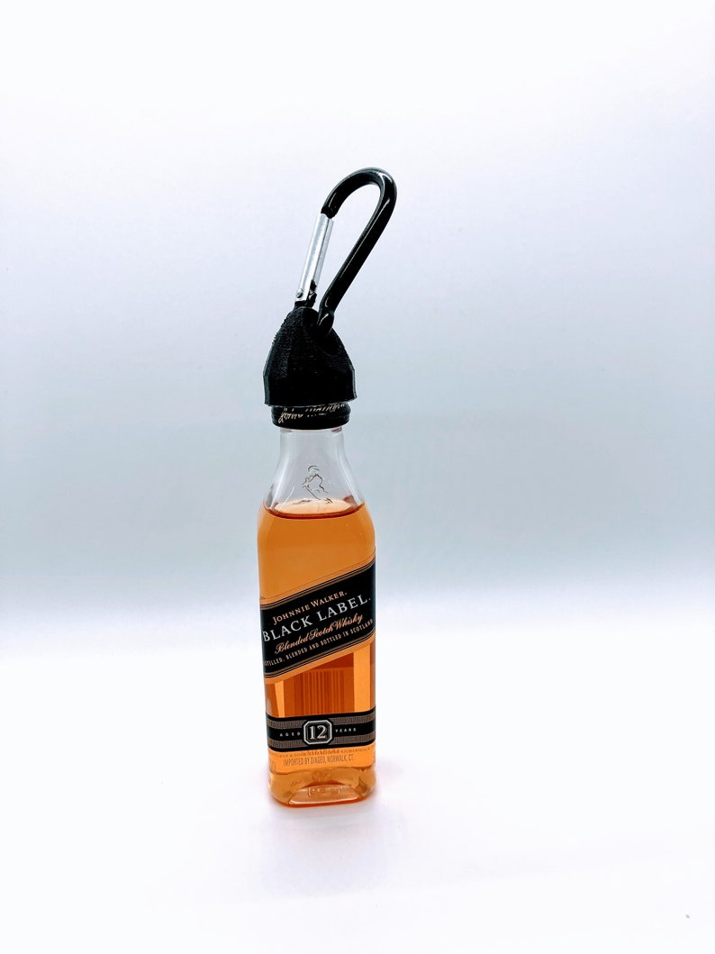 May include: A miniature bottle of Johnnie Walker Black Label Blended Scotch Whisky with a black carabiner attached to the top. The bottle is clear glass and the label is black with gold lettering. The label reads "Johnnie Walker Black Label Blended Scotch Whisky" and "12 Years Old".