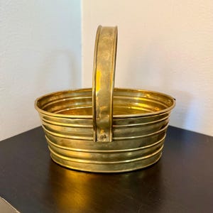 May include: An oval-shaped brass basket featuring a polished metallic finish and horizontal ribbed detailing along the exterior walls. The basket includes a single upright handle attached to the center of the sides with small metal rivets.
