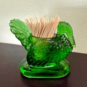 May include: A vibrant green glass toothpick holder shaped like a turkey with detailed moulded feathers and a curved neck. The translucent glass container is filled with a cluster of light-coloured wooden toothpicks, standing on an oval base.