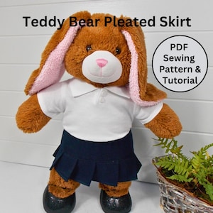 Può includere: Un orsacchiotto di peluche marrone che indossa una camicia bianca con colletto e una gonna a pieghe blu navy. L'orso ha orecchie di coniglio rosa. Il testo "PDF Sewing Pattern & Tutorial" è in un cerchio sopra l'orso.