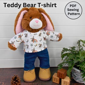 May include: A brown teddy bear wearing a white long-sleeved t-shirt with a rainbow and bunny print, blue jeans, and brown shoes. The bear is standing on a wooden floor with wooden blocks and a potted plant in the background. The text "PDF Sewing Pattern" is visible in the top right corner of the image.