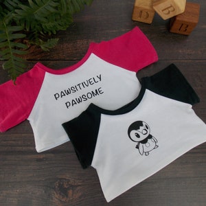 May include: Two white t-shirts with different designs. One has black sleeves and a penguin graphic, the other has pink sleeves and the text "Pawsitively Pawsome".