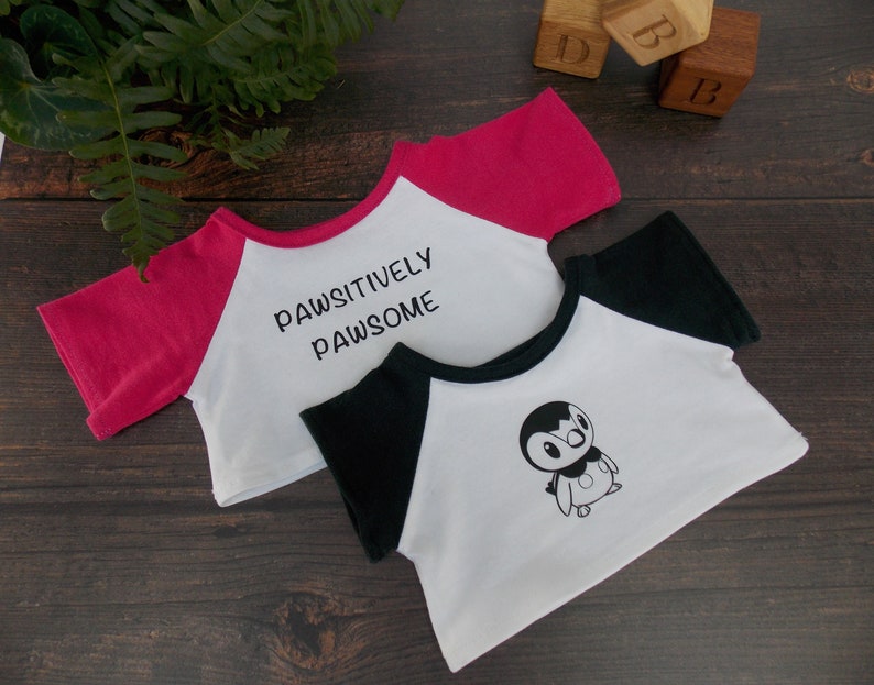 May include: Two white t-shirts with different designs. One has black sleeves and a penguin graphic, the other has pink sleeves and the text "Pawsitively Pawsome".