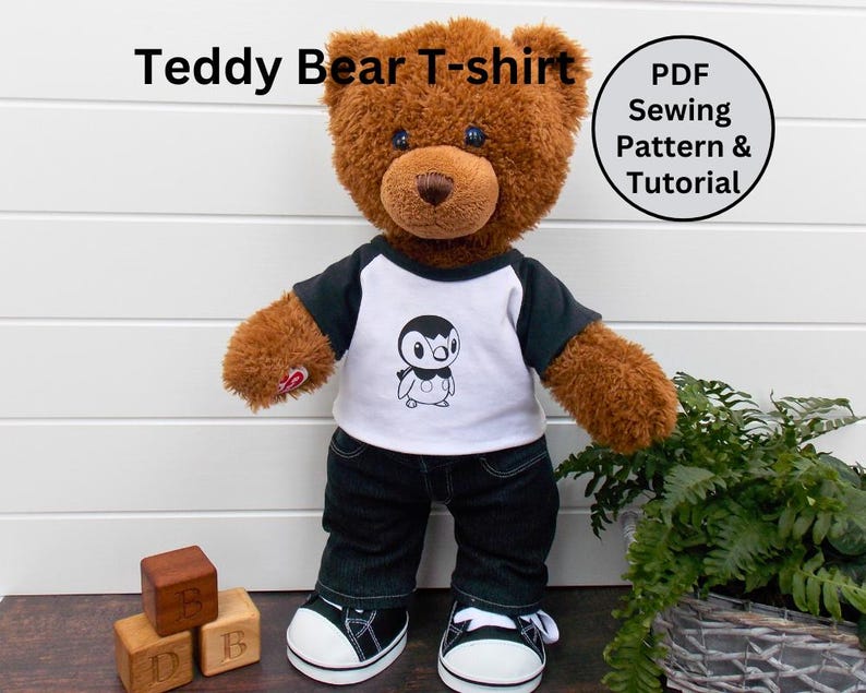 May include: A brown teddy bear wearing a white t-shirt with a black penguin graphic and black jeans. The t-shirt has a black and white ringer design. The bear is standing on a wooden surface with wooden blocks in the foreground. The text "Teddy Bear T-shirt" is in the top left corner of the image. The text "PDF Sewing Pattern & Tutorial" is in a circle in the top right corner of the image.