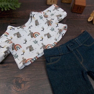 May include: A white short-sleeved shirt with a pattern of brown rabbits and colorful rainbows. The shirt is paired with dark blue denim pants.