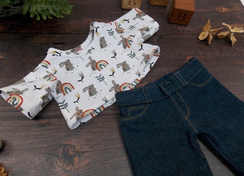 May include: A white short-sleeved shirt with a pattern of brown rabbits and colorful rainbows. The shirt is paired with dark blue denim pants.