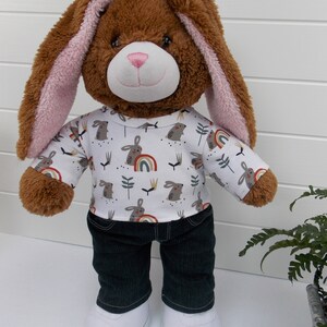 Build a bear teddy bear wearing a white t-shirt with rabbits and rainbows on, black jeans and white trainers.