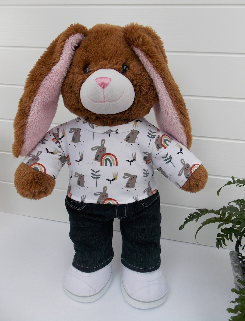 Build a bear teddy bear wearing a white t-shirt with rabbits and rainbows on, black jeans and white trainers.