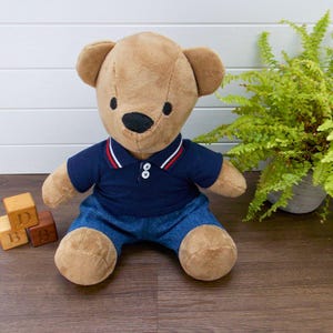 May include: A plush teddy bear wearing a navy blue polo shirt with red and white trim and blue denim pants. The bear has a black nose and small black eyes. Wooden alphabet blocks are visible in the lower left corner.