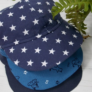 Pu&ograve; includere: Tre cappellini da baseball blu e bianchi con motivi impilati uno sopra l'altro. Il cappellino superiore ha uno sfondo blu scuro con stelle bianche. Il cappellino intermedio ha uno sfondo blu chiaro con impronte di zampe bianche e un disegno di un cane. Il cappellino inferiore &egrave; di un blu scuro pieno.