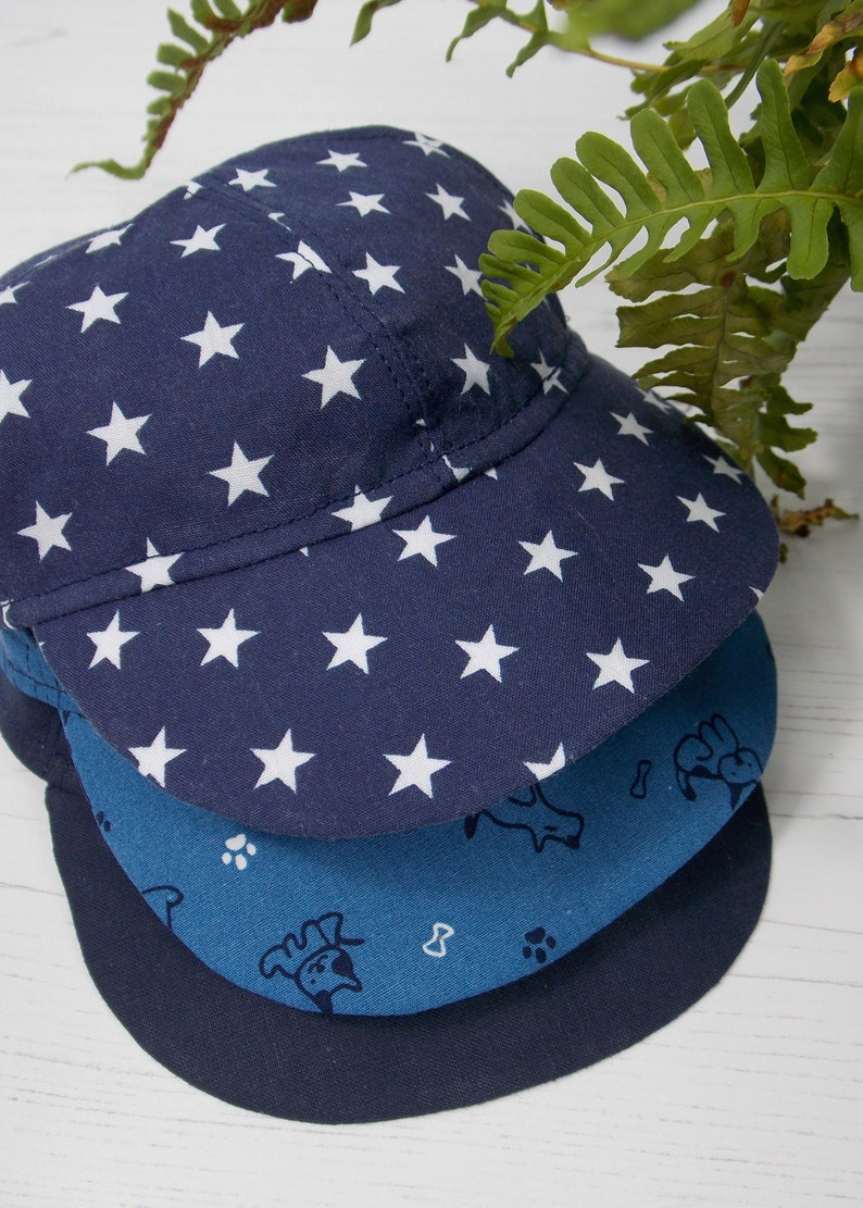 Pu&ograve; includere: Tre cappellini da baseball blu e bianchi con motivi impilati uno sopra l'altro. Il cappellino superiore ha uno sfondo blu scuro con stelle bianche. Il cappellino intermedio ha uno sfondo blu chiaro con impronte di zampe bianche e un disegno di un cane. Il cappellino inferiore &egrave; di un blu scuro pieno.
