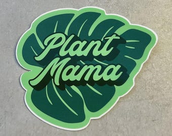 May include: A green sticker in the shape of a leaf with the words "Plant Mama" in a cursive font. The leaf is a darker green with lighter green accents and a white border. The sticker is on a light gray surface.