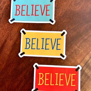 以下が含まれることがあります： 太字の「BELIEVE」という言葉が入った長方形のステッカーが3枚。ステッカーの色は、水色、黄色、赤と様々です。各ステッカーには白い縁取りと黒い角のアクセントが付いています。
