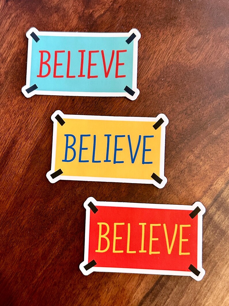 以下が含まれることがあります： 太字の「BELIEVE」という言葉が入った長方形のステッカーが3枚。ステッカーの色は、水色、黄色、赤と様々です。各ステッカーには白い縁取りと黒い角のアクセントが付いています。