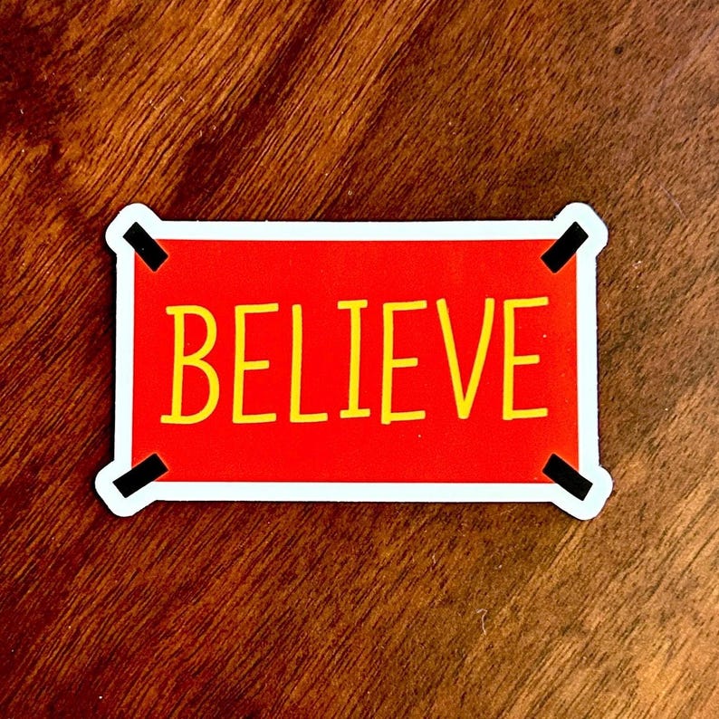 以下が含まれることがあります： 赤色の背景に、黒い縁取りの黄色い文字で「BELIEVE」と書かれた長方形のステッカー。ステッカーには白い縁と黒い角のアクセントがあります。ステッカーは木製の表面にあります。