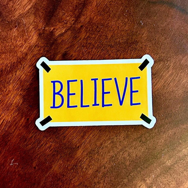 以下が含まれることがあります： 黄色い背景に青い大文字で「BELIEVE」と書かれた長方形のステッカー。ステッカーは白い縁取りと黒い角のアクセントがあり、茶色の木製の表面に配置されています。