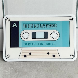 May include: A white sticker in the shape of a cassette tape. The tape has a light blue center with the text "THE BEST MIX TAPE EVERRRRR" and "RETRO LOVE NOTES". The left side is dark blue with a white "A".