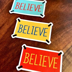 以下が含まれることがあります： 太字の「BELIEVE」という言葉が入った長方形のステッカー3枚。ステッカーの色は、赤字のライトブルー、青字のイエロー、黄字のレッドと様々です。各ステッカーには黒いコーナーアクセントが付いています。