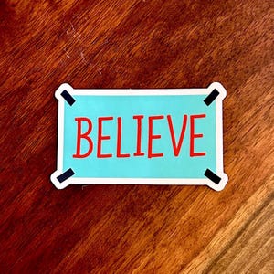 以下が含まれることがあります： 水色の背景に赤い文字で「BELIEVE」と書かれた長方形のステッカー。白い縁取りと黒い角のアクセントがあり、木製の表面に配置されています。