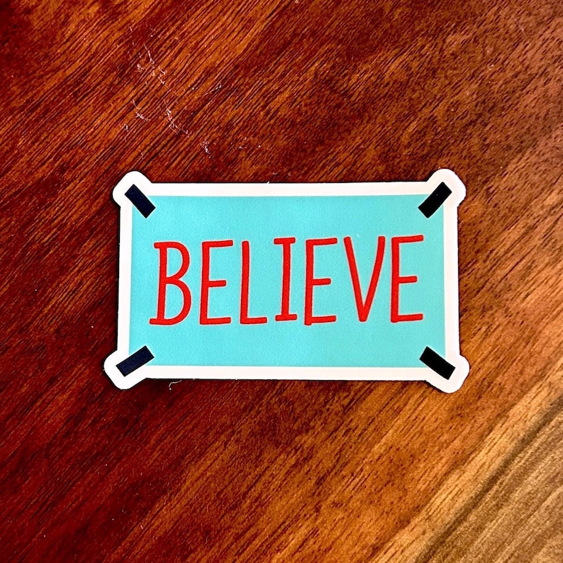 以下が含まれることがあります： 水色の背景に赤い文字で「BELIEVE」と書かれた長方形のステッカー。白い縁取りと黒い角のアクセントがあり、木製の表面に配置されています。