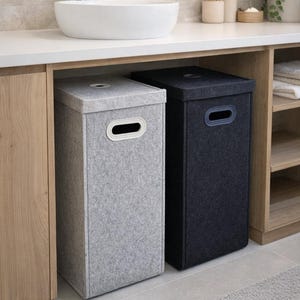 May include: Two rectangular laundry hampers, one light gray and one dark gray, sit side-by-side under a countertop. Each hamper has a lid with a circular opening and a handle. The hampers are made of a felt-like material and are in a bathroom setting.