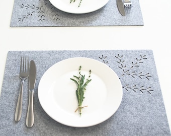 May include: Two rectangular light gray felt placemats featuring a laser-cut branch pattern in the corner. Each placemat holds a white ceramic plate topped with a sprig of green herbs, a stainless steel fork, and a stainless steel knife.