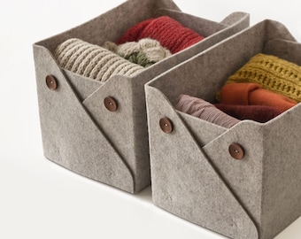 May include: Two rectangular light grey felt storage bins with folded side flaps secured by small brown wooden buttons. The bins are filled with folded knit garments in various colours including red, beige, mustard yellow, and orange.