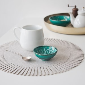 May include: A white mug, a teal bowl with a decorative design, and a silver spoon on a grey felt coaster with a fringe edge.