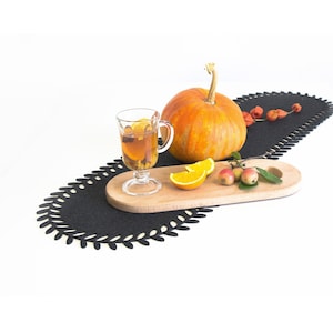 May include: A black felt table runner with a decorative leaf pattern, a wooden serving tray with a glass of cider, orange slices, and small red apples, and a whole orange pumpkin.