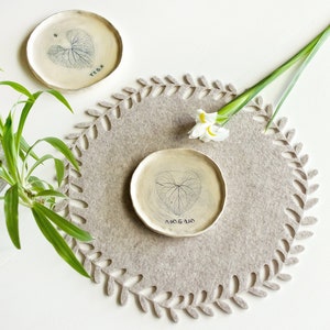 May include: A beige felt placemat with a decorative leaf pattern around the edge. Two small white ceramic plates with leaf designs and the words "YOGA" and "AROMA" are on the placemat.