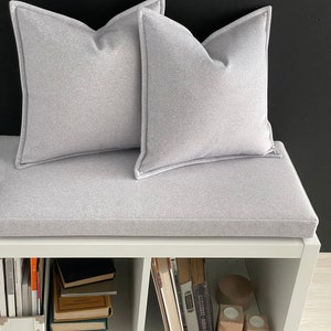 Op de afbeelding: Een witte bank met een grijs kussen en twee grijze kussens. De bank heeft drie planken, die elk gevuld zijn met boeken. De planken zijn wit en de boeken zijn in verschillende kleuren.