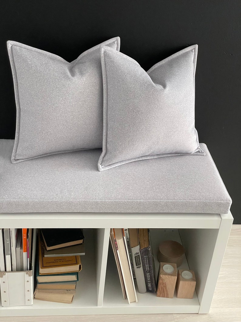 Op de afbeelding: Een witte bank met een grijs kussen en twee grijze kussens. De bank heeft drie planken, die elk gevuld zijn met boeken. De planken zijn wit en de boeken zijn in verschillende kleuren.