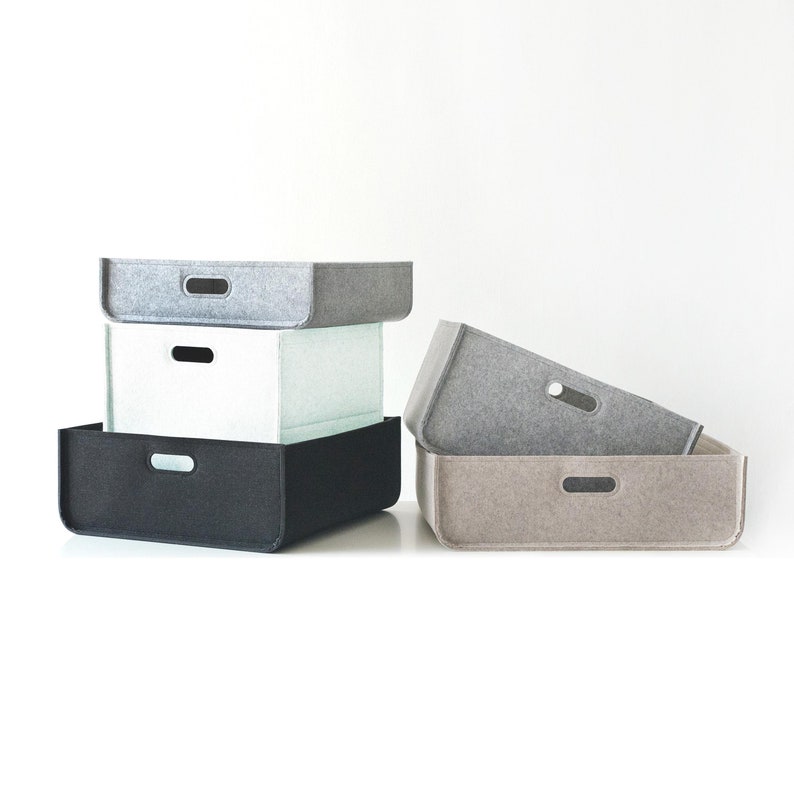 May include: A stack of four rectangular storage bins in various colors: black, light gray, white, and tan. Each bin has a rounded edge and a small, oval-shaped handle cut out on the front. The bins are made of a felt-like material.