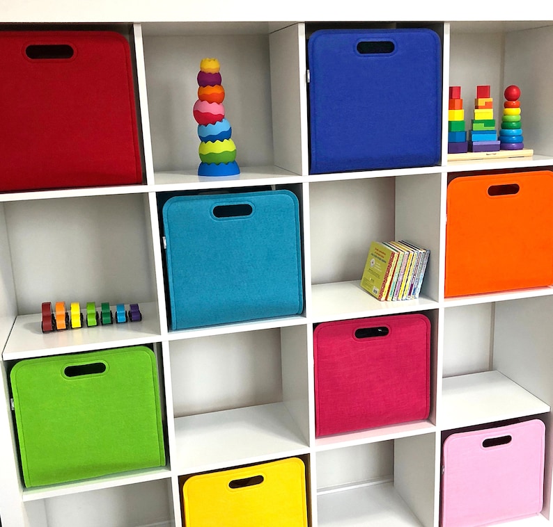 May include: A white shelving unit filled with colorful storage boxes and children's toys. The boxes are red, blue, turquoise, orange, green, pink, and yellow. Toys include a stacking tower, wooden blocks, and a set of small books.