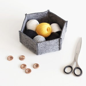 Puede incluir: Una cesta de almacenamiento hexagonal de fieltro gris llena de ovillos de lana en blanco, amarillo y negro. También hay botones de madera y un par de tijeras plateadas con mangos negros sobre una superficie blanca.