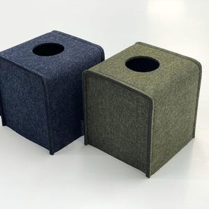 May include: Two square tissue box covers. One is navy blue, the other is olive green. Both have a rounded top with a circular opening and a small black tag on the side. The boxes are made of a felt-like material.