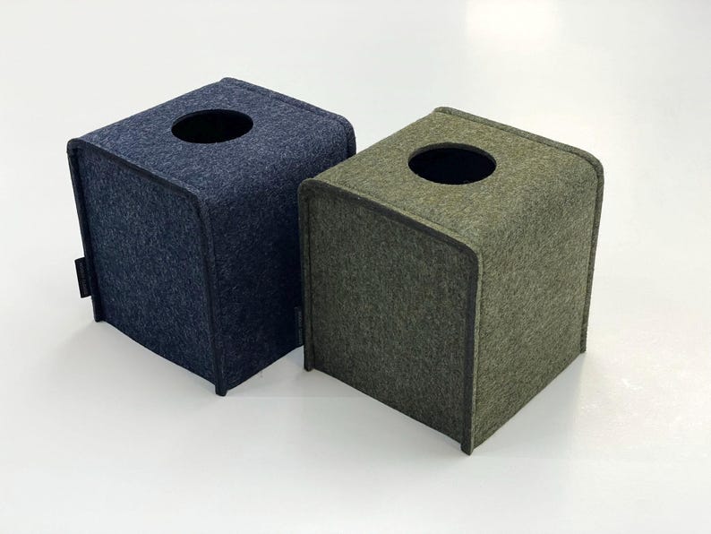 May include: Two square tissue box covers. One is navy blue, the other is olive green. Both have a rounded top with a circular opening and a small black tag on the side. The boxes are made of a felt-like material.