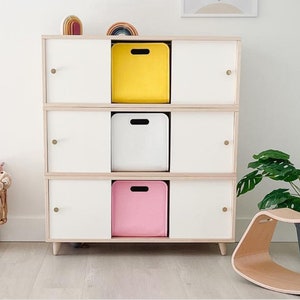 May include: A three-tiered white and wood-toned storage cabinet with three cubbies, each holding a yellow, white, and pink bin. A wooden rocking toy and a potted plant are nearby. A small wicker basket with stuffed animals is on the left.