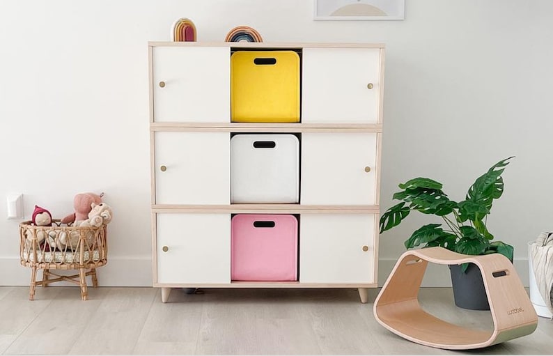 May include: A three-tiered white and wood-toned storage cabinet with three cubbies, each holding a yellow, white, and pink bin. A wooden rocking toy and a potted plant are nearby. A small wicker basket with stuffed animals is on the left.