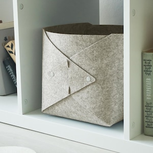 May include: A light gray felt storage bin with a folded, envelope-style front secured by two small circular fasteners, positioned on a white shelf between two stacks of hardcover books with various titles in different languages and scripts.