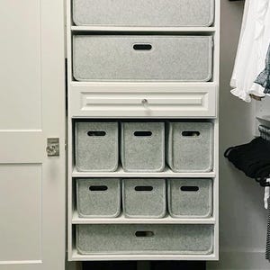 May include: A white closet organizer with multiple shelves and drawers. The shelves contain gray felt storage bins of various sizes, including rectangular and square shapes. The closet door is white, and the overall aesthetic is clean and organized.