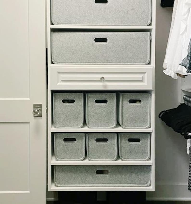 May include: A white closet organizer with multiple shelves and drawers. The shelves contain gray felt storage bins of various sizes, including rectangular and square shapes. The closet door is white, and the overall aesthetic is clean and organized.