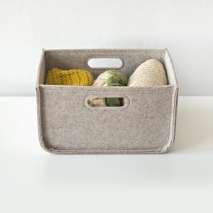 May include: A rectangular, light gray felt storage bin with two handle cutouts. The bin contains three knitted items in yellow, green, and cream colors. The bin is sitting on a white surface.