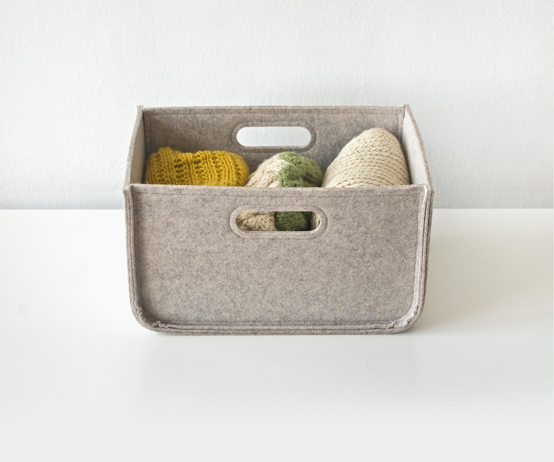 May include: A rectangular, light gray felt storage bin with two handle cutouts. The bin contains three knitted items in yellow, green, and cream colors. The bin is sitting on a white surface.