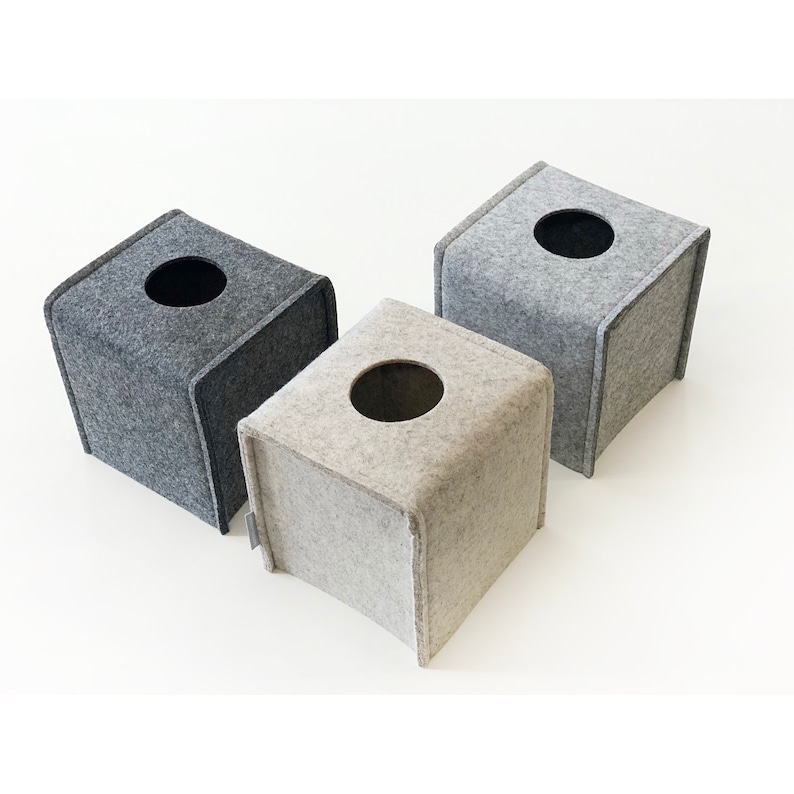 May include: Three square tissue box covers in shades of gray. Each box has a circular opening on top. The boxes are made of a soft, felt-like material. The boxes are arranged on a white surface.