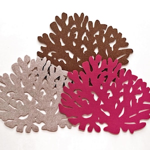 Op de afbeelding: Drie vilten onderzetters in koraalvorm in bruin, taupe en magenta tinten. De onderzetters hebben een gedetailleerd, organisch ontwerp met uitgesneden patronen. Ze zijn gerangschikt op een wit oppervlak, waarbij hun kleuren en texturen worden getoond.