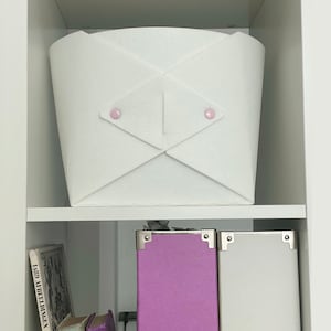 May include: White felt storage basket with pink buttons on the front. The basket is sitting on a white shelf.